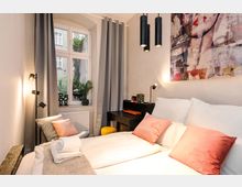 Modernes Schlafzimmer mit Doppelbett, weißen Bettdecken und rosafarbenen Kissen, dazu graue Vorhänge und ein großes Fenster mit Blick in einen begrünten Innenhof. Im Raum stehen ein schwarzer Schreibtisch mit Hocker, mehrere schwarze Lampen und ein großes Wandbild über dem Bett.