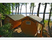 Zwei kleine, runde Holzhütten mit grün gedeckten Dächern stehen in einem waldigen Bereich am Ufer eines Sees. Im Hintergrund sind Wasser und Bäume am gegenüberliegenden Ufer sichtbar.