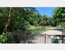 Metallzaun mit geöffnetem Tor führt in einen Park mit Wegen, Blumenbeeten und dichtem Baumbestand. Im Hintergrund sind Wiesen, rote Blumen und zwei Spaziergänger sichtbar.