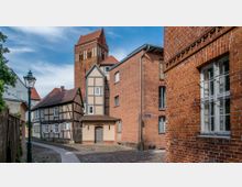 Gepflegte Altstadtgasse mit Kopfsteinpflaster, Fachwerkhäusern und roten Ziegelgebäuden; im Hintergrund ragt ein historischer Kirchturm aus roten Backsteinen hervor. Links steht eine grüne Straßenlaterne, die von Bäumen und Holzzäunen gesäumt wird.