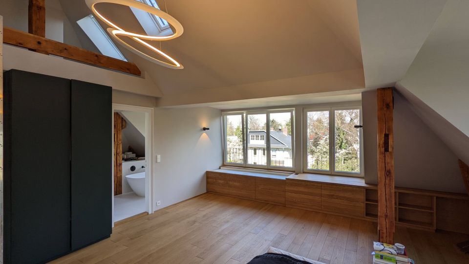 Modernes Dachgeschosszimmer mit schrägen Decken, sichtbaren Holzbalken und hellem Holzboden, beleuchtet durch große Fensterfront und ein Dachfenster. Unter den Fenstern verläuft eine lange Holzkommode; links steht ein hoher, dunkler Schrank und daneben führt eine Tür in ein Badezimmer.