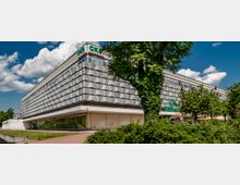 Modernes Einkaufszentrum mit grauer, geometrisch strukturierter Fassade, einem „CKS" Schild und umliegender Begrünung. Im Vordergrund sind ein gepflasterter Gehweg, Sträucher und ein Baum sichtbar, unter einem sonnigen Himmel.