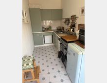 Schmale Küche mit mintfarbenen Schränken, weißen Fliesen an der Wand und gemustertem Boden. Links befinden sich Arbeitsflächen mit Spüle und Geräten, rechts ein Retro-Kühlschrank, ein Holzregal und ein freistehender Backofen.