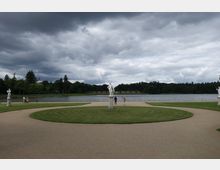 Ein symmetrischer Park mit einem zentralen, kreisförmigen Weg, umgeben von gepflegtem Rasen. In der Mitte steht eine weiße Statue auf einem Sockel, im Hintergrund ein See mit umgebenden Bäumen unter bewölktem Himmel.