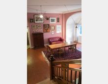 Wohnzimmer mit Holztreppe und klassischer Einrichtung; im Zentrum steht ein langer Holztisch mit Stühlen auf einem roten Teppich, dahinter ein rotes Ledersofa vor einer rosa Wand, die mit Kunstwerken und Bildern dekoriert ist. Ein großer heller Durchgang führt zu einem weiteren Bereich mit Fenster.