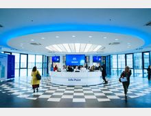 Großer, moderner Empfangsbereich mit einem ovalen Info-Point in der Mitte, flankiert von Mitarbeitern hinter der Theke. Der Raum hat eine schwarz-weiß karierte Bodenfläche, eine gläserne Außenfassade und eine beleuchtete Deckeninstallation mit blauer Akzentbeleuchtung.