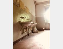 Ein verlassener Waschraum mit mehreren an der Wand montierten Waschbecken ohne Wasserhähne, sichtbaren Rohrleitungen und abblätternder Wandfarbe. Im Hintergrund fällt Licht durch ein Fenster mit beschädigtem Rahmen auf einen teilweise verschmutzten Boden.