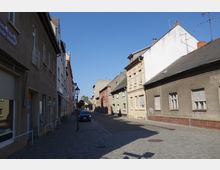 Schmale Straße in einem Wohngebiet mit älteren Gebäuden auf beiden Seiten, teilweise renoviert, mit hellen und erdigen Fassadenfarben. Kopfsteinpflaster bedeckt den Gehweg, während vereinzelt Laternen und ein geparktes Auto sichtbar sind; der Himmel ist klar und blau.