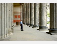 Säulenhalle eines historischen Gebäudes mit großen, massiven Steinsäulen und einer dekorativen Rückwand in rot-beigen Farben; ein Mann in Uniform steht in der Halle.