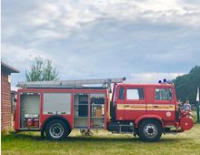 Ein rotes Feuerwehrfahrzeug mit der Aufschrift „Räddningstjänsten“ steht auf einer Wiese. Die Fahrzeugseite ist geöffnet und zeigt Ausrüstung wie einen Generator und Werkzeuge, im Hintergrund sind Bäume und ein bewölkter Himmel sichtbar.