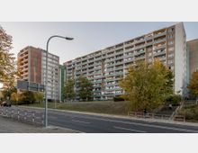 Mehrstöckiger Plattenbau mit Balkonen und farbigen Fassadenelementen, umgeben von Bäumen und Grünflächen. Im Vordergrund führt eine Straße mit einem Fußgängerweg und Straßenschildern am linken Bildrand vorbei.