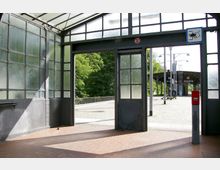 Ein Wartebereich eines Bahnhofs mit verglasten Metallwänden und einem Glasdach, das natürliches Licht durchlässt. Im Hintergrund ist ein gepflasterter Bahnsteig mit Sitzbänken, Laternen und einem Vordach zu sehen; daneben ein roter Briefkasten und Hinweisschilder.