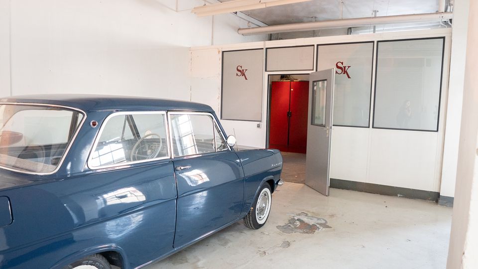Innenraum einer hellen Werkstatt- oder Garagenhalle mit weiß gestrichenen Wänden, sichtbaren Deckenrohren und abgenutztem Betonboden. Links steht ein dunkelblauer Oldtimer, rechts sind verglaste Trennwände mit einer offenen Tür zu einem roten Nebenraum zu sehen.