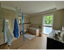 Helles Badezimmer mit Holzbodenoptik, einem freistehenden Waschbecken und einer Badewanne. Ein Fenster mit Blick ins Grüne sorgt für natürliches Licht, während Handtücher, Pflanzen und Körbe die Einrichtung ergänzen.