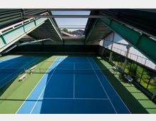 Innenansicht einer Tennishalle mit zwei Tennisplätzen in Blau und Grün, umgeben von hohen Fenstern, die Tageslicht hereinlassen. Die Halle hat eine Metallkonstruktion mit sichtbaren Trägern und ein teilweise lichtdurchlässiges Dach.