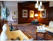 Wohnzimmer mit Holzwänden und Teppichboden, eingerichtet mit einer cremefarbenen Couch, einem schwarzen Liegesessel und modernen Hängelampen. An der Wand hängen gerahmte Kunstwerke, im Raum stehen ein großer Kaktus und ein kleiner Esstisch im hinteren Bereich.