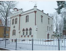 Zweistöckiges historisches Gebäude mit weißer Fassade und Ziegelverzierungen, umgeben von Schnee. Im Vordergrund befindet sich ein Metallzaun, dahinter Bäume im winterlichen Ambiente.