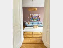 Blick durch eine weiße Flügeltür in ein helles Wohnzimmer mit Parkettboden, großem hellem Teppich, grauem Sofa und einem Glas-Couchtisch. An der roséfarbenen Wand hängt ein großes, farbenfrohes Kunstwerk; darüber eine Deckenstuckleiste und eine runde Pendelleuchte.
