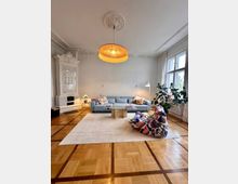 Wohnzimmer mit hellem Parkettboden, einer großen blauen Sofalandschaft, einem rechteckigen Glastisch und einer Sitzgelegenheit mit buntem Stoffbezug. Im Hintergrund befinden sich ein dekorativer weißer Kachelofen, eine Hängelampe mit geflochtenem Lampenschirm und ein großes Fenster mit weißen Vorhängen.