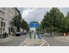 Eingang zur U-Bahn-Station "Rosa-Luxemburg-Platz" mit einem blauen Hinweisschild, umgeben von Bäumen und flankiert von mehrstöckigen Gebäuden auf der linken Seite. Die Umgebung zeigt eine Straße mit parkenden Autos und Gehwegen.