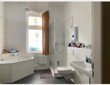 Helles Badezimmer mit weißen Wandfliesen und dunklem Boden, ausgestattet mit Eckbadewanne, verglaster Dusche, wandhängendem WC und großem Waschbecken. Ein großes Fenster mit Vorhang bringt Tageslicht hinein; auf einer Ablage stehen Pflegeprodukte und eine Pflanze.