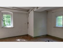 Ein leerer Raum mit zwei vergitterten Fenstern, die nach draußen ins Grüne blicken. Die Wände sind einfach verputzt, die Decke hat sichtbare Rohre, und der Boden ist mit abgenutzten Fliesen bedeckt.