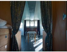 Innenansicht eines Bootsraums mit Blick auf einen Schiffsteuerstand. Im Vordergrund sind schwere Vorhänge und Holzmöbel zu sehen; dahinter ein Tisch mit Stühlen vor dem Fenster und einem Steuerrad.