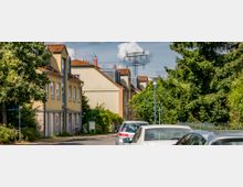 Straßenszene in einem Wohngebiet mit gelb verputzten Reihenhäusern und roten Ziegeldächern, umgeben von Bäumen und Sträuchern. Geparkte Autos stehen entlang der Straße, im Hintergrund ist ein Strommast sichtbar.