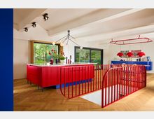 Moderne Küche mit offenen Wohnraum-Elementen, dominiert von einer großen roten Kücheninsel mit weißen Arbeitsflächen. Auffällige rote Geländer um eine Treppenöffnung, Parkettboden in Fischgrätmuster und große Fenster mit Blick auf eine grüne Außenlandschaft prägen den Raum.