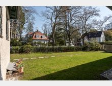 Gepflegter Garten mit grünem Rasen, gesäumt von einem niedrigen Holzzaun und Büschen. Im Hintergrund sind einzelne Einfamilienhäuser mit roten und dunklen Dächern sowie hohe Bäume sichtbar, die einen ruhigen Wohnviertelcharakter vermitteln.