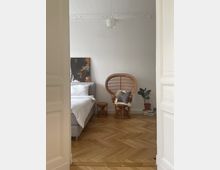 Ein Schlafzimmer mit hellen Wänden und Fischgrätparkettboden. Links steht ein grau bezogenes Bett mit weißer Bettwäsche, rechts ein Korbsessel mit Kissen und Fell, daneben ein kleiner runder Beistelltisch und eine Zimmerpflanze. An der Decke ist eine dekorative Lampe angebracht.