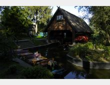 Holzgebäude mit der Aufschrift „Bootshaus Conrad“ neben einem schmalen Kanal, umgeben von Bäumen und Grünflächen. Im Vordergrund sind Tische und Bänke mit karierten Decken direkt am Wasser sowie kleine Boote sichtbar.