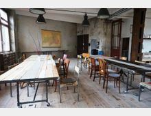 Ein rustikaler Raum mit Holzboden und mehreren langen Holztischen auf Metallgestellen, umgeben von unterschiedlichen Stühlen. Im Hintergrund sind eine beigen Wand mit einer angehefteten Karte, große Industrie-Lampen und alte Holztüren sichtbar.