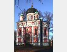 Eine orthodoxe Kirche mit einer großen zentralen Kuppel, flankiert von vier kleineren grünen Kuppeln. Die Fassade ist in Rot- und Weißtönen gehalten, mit Bögen und dekorativen Elementen, umgeben von kahlen Bäumen und einem niedrigen Zaun.