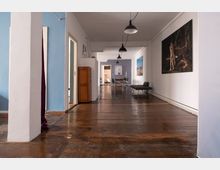 Langer Flur in einem Innenraum mit Holzboden, weißen Wänden und blauen Akzenten; an den Wänden hängen große Kunstwerke, die von Deckenlampen beleuchtet werden. Am Ende des Flurs sind ein Esstisch mit Stühlen und eine Couch sichtbar.