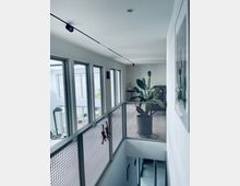 Moderner Innenraum mit einem hellen Flur, der von bodentiefen Fenstern auf der linken Seite beleuchtet wird. Der Flur verfügt über ein Geländer aus Metallgitter, eine große Topfpflanze (Kaktus) am Ende des Gangs und eine minimalistische Deckenbeleuchtung.