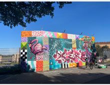 Eine bunte Wand mit großflächigem Graffiti, darunter ein rosa Flamingo, ein abstrakter Wal und Schriftzüge in lebhaften Farben. Im Vordergrund sind ein Fahrrad und ein Elektro-Scooter abgestellt, während im Hintergrund ein umzäuntes Gelände und einige Gebäude sichtbar sind.