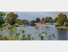 Flusslandschaft mit kleiner Marina, mehreren geparkten Booten und einem roten Gebäude mit Satteldach im Hintergrund. Auf dem Wasser fährt ein kleines Boot, umgeben von Bäumen und Grünflächen.