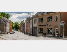 Straßenszene in einer Kleinstadt mit zweigeschossigen Gebäuden, darunter ein beiges Haus mit der Aufschrift "Seniorenbetreuung WG", und geparkten Autos entlang der Straße. Im Hintergrund sind Bäume und Wolken am Himmel zu sehen.