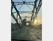 Eine Stahlfachwerkbrücke bei Sonnenuntergang, aufgenommen aus einem Fahrzeug. Im Hintergrund sind ein mehrstöckiges Gebäude und Bäume sichtbar, während die Brücke den Himmel dominiert.