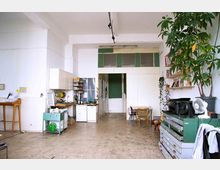 Ein heller, offener Wohnraum mit einer kleinen Küchenzeile links, einem Tisch mit Stühlen im Hintergrund und Regalen an der rechten Wand. Der Raum hat hohe Decken, eine Zimmerpflanze und eine Mischung aus funktionalen und dekorativen Möbeln.