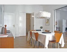 Esszimmer in einem hellen, modernen Interieur mit Holzboden, einem Rechtecktisch mit weißer Tischdecke, sechs Holzstühlen und einer auffälligen weißen Hängelampe. Im Hintergrund sind eine Kommode mit Dekoration, weiße Türen und minimalistische Wandkunst zu sehen.