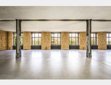 Großer, leerer Loft- oder Industriehallenraum mit glattem Betonboden, sichtbaren Stahlstützen und einer langen Backsteinwand. Mehrere hohe Sprossenfenster mit Heizkörpern darunter lassen viel Tageslicht in den Raum.