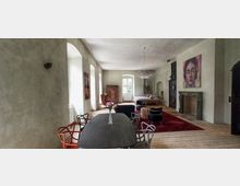 Innenraum eines stilvollen Wohnbereichs mit hellen Wänden, einem Holzboden und einem großen Tisch mit unterschiedlichen Stühlen im Vordergrund. Im Hintergrund befinden sich ein Bett, ein roter Teppich, ein Kamin mit grüner Umrahmung sowie Wandkunst und ein Kronleuchter.