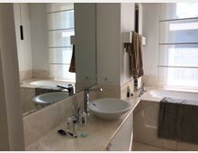 Modernes Badezimmer mit einer großen Spiegelwand über einem Waschtisch mit Aufsatzwaschbecken. Rechts befindet sich eine freistehende Badewanne mit hellen Marmorelementen und halbtransparenten Rollläden an den Fenstern.