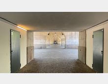 Blick durch einen überdachten Gang mit einer Reihe von Rundbögen aus Stein, flankiert von Wänden und Türen. Der Boden ist mit Kopfsteinpflaster bedeckt, und in der Mitte hängen gleichmäßig verteilte Lampen.