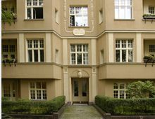 Fassade eines historischen Wohnhauses mit hellbeigem Putz und verzierten Fensterelementen. Der Eingang ist von symmetrisch angeordneten Fenstern sowie dezenten Grünflächen mit Hecken eingefasst, darüber ein Schild mit der Jahreszahl "1912–1913".