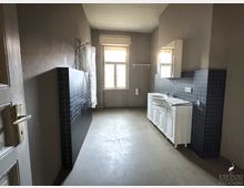 Ein schlichter Raum mit grauen Wänden und einem Fenster, das Tageslicht hereinlässt. Der Raum enthält eine Dusche mit schwarzer Fliesenwand, ein weißes Waschbecken auf einem Unterschrank und einen Spiegel an der Wand.