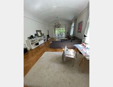 Wohnzimmer mit hellem Parkettboden, großem Teppich und einer grauen Sofalandschaft. Im Hintergrund führt ein offener Durchgang zu einem Essbereich mit Blick auf eine grüne Terrasse; eine bunte Hängelampe und hohe Fenster sorgen für viel Tageslicht.