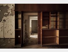 Ein verlassener Raum mit stark abblätternder Farbe an den Wänden, einem Holzregal mit offenen Fächern und einem Türdurchgang, der in einen weiteren, ebenfalls verfallenen Raum führt. Der Boden ist mit Schmutz und Trümmern bedeckt, die Umgebung wirkt stark verfallen und ungepflegt.